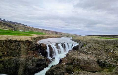       Small but powerful waterfall plunging into a rocky gorge amid Icelandic highlands with two distant visitors for scale.
  
