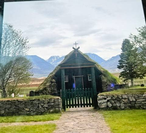       Grass-roofed wooden church set against snowy mountains with spring foliage in the foreground.
  