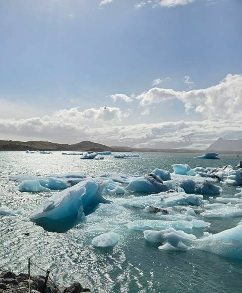       Icebergs drifting across a shimmering glacial lagoon under scattered clouds and distant snow-capped peaks.
  