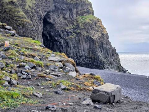       Dramatic basalt column cliff and cave opening at a black-sand beach beside the shimmering sea.
  