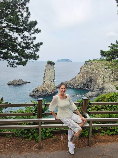       A smiling visitor stands before a dramatic sea stack and turquoise cove on Jeju Island.
  