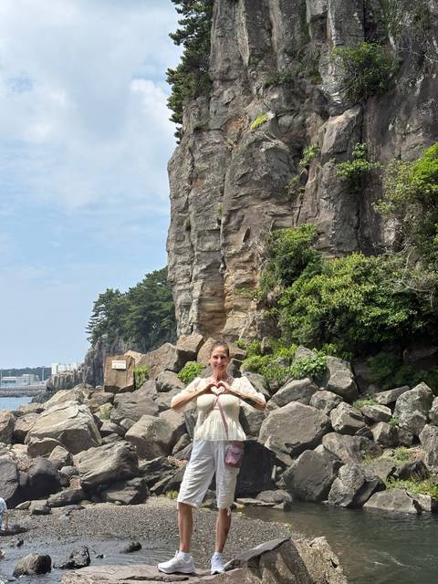       A woman forms a heart with her hands in front of rugged seaside cliffs and boulders.
  