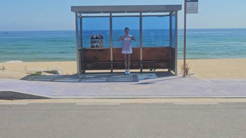       A lone traveler poses inside a glass bus stop shelter facing a wide sandy beach and blue sea.
  