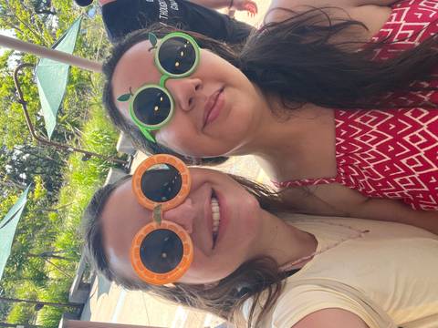       Two women smile wearing playful fruit-themed sunglasses in bright outdoor light.
  