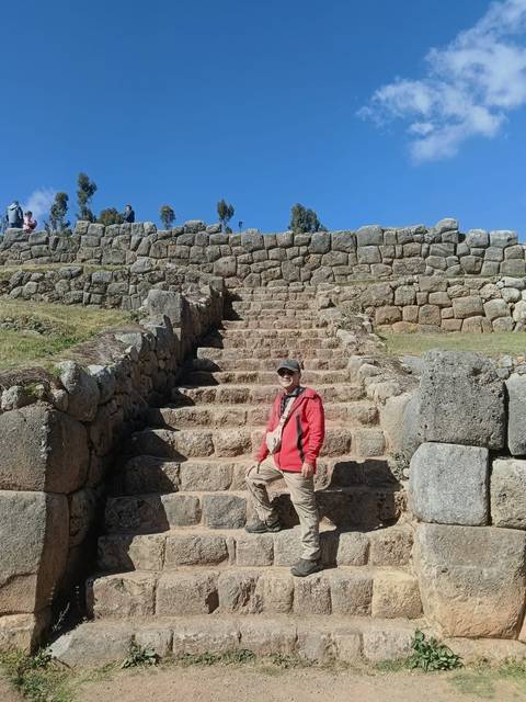       Visitor poses on ancient stone steps of Inca ruins with massive terrace walls under clear skies.
  