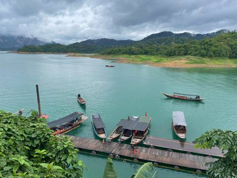       Long-tail boats moored on emerald green lake surrounded by jungle-covered limestone hills under cloudy skies.
  