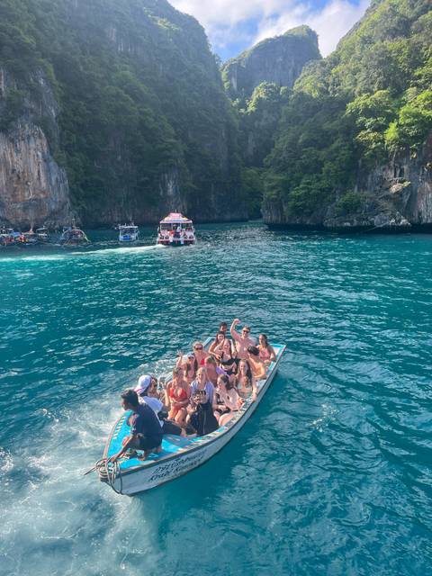       Group of cheerful travellers waving from a small boat on bright turquoise water with steep cliffs behind.
  
