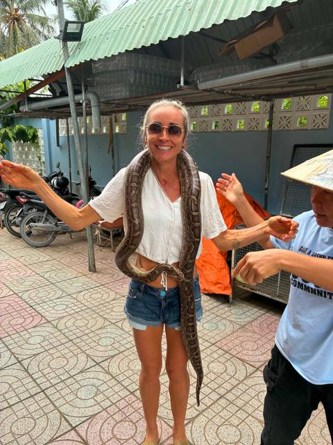       Traveller smiling while a large snake is draped around her shoulders, assisted by a local handler.
  