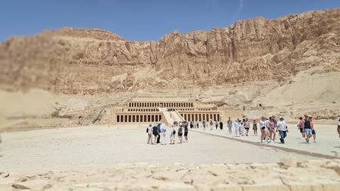       Temple of Hatshepsut dwarfed by sheer cliffs with visitors walking the grand causeway.
  