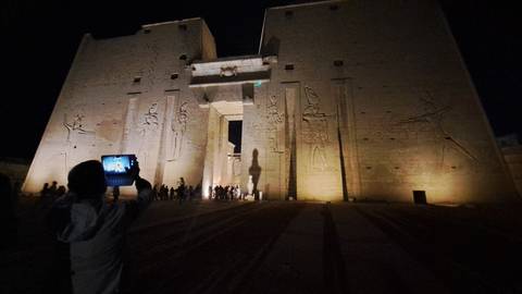       Night view of Edfu Temple illuminated with tourists capturing photos at the entrance.
  
