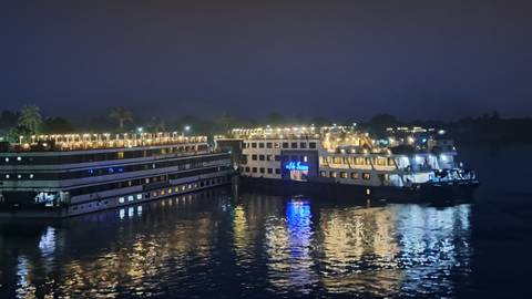       Nile cruise ships moored side by side, deck lights reflecting on the dark river at night.
  