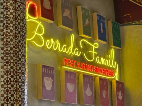       Neon sign reading 'Berrada Family Restaurant Since 1999' set against tiled pillar and framed posters.
  