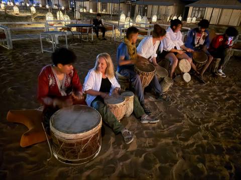       Nighttime drum circle on desert sand with motion-blurred participants playing hand drums.
  