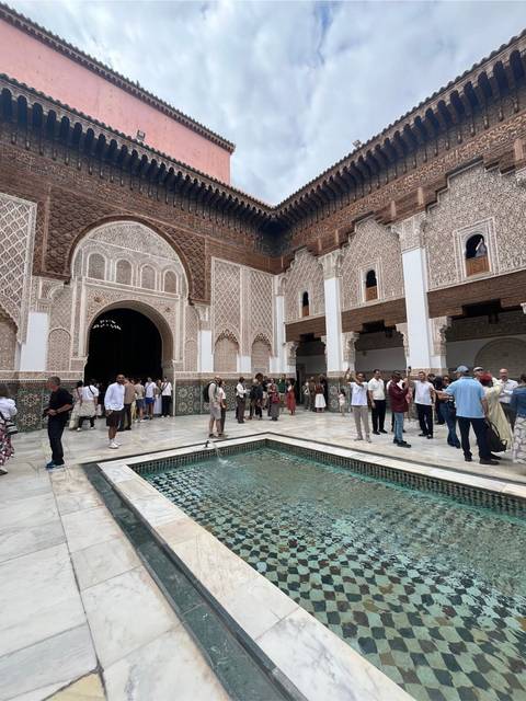       Visitors explore ornate courtyard of a Moroccan madrasa featuring carved stucco and a central pool.
  