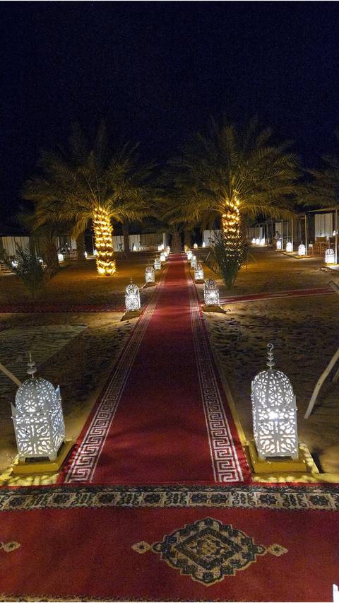       Nighttime desert camp walkway lined with glowing lanterns and palm trees
  