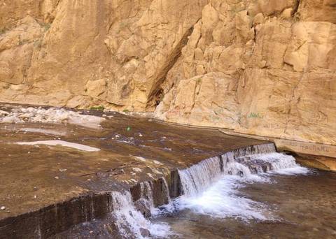       Small cascade flowing over a ledge into a shallow rocky stream beneath tan cliffs
  