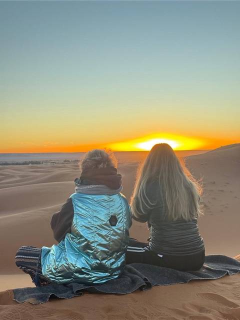       Two women sit on a dune watching the sun dip below the Sahara horizon
  
