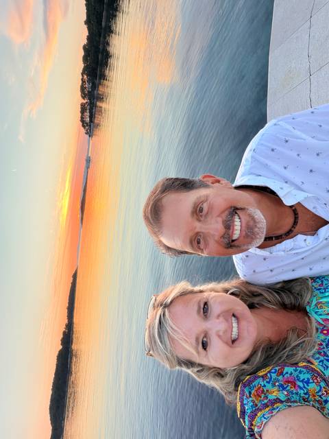       Smiling couple selfie aboard a boat with a vivid orange sunset reflecting on calm sea.
  