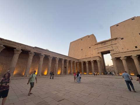      Interior court of Edfu Temple with columns lit by uplights while visitors wander during twilight.
  