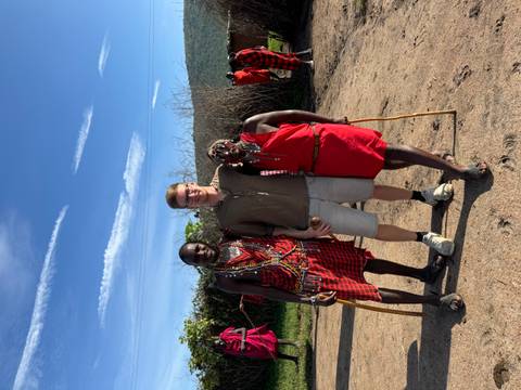       Traveler poses with two Maasai men dressed in traditional red shukas against a bright blue sky.
  