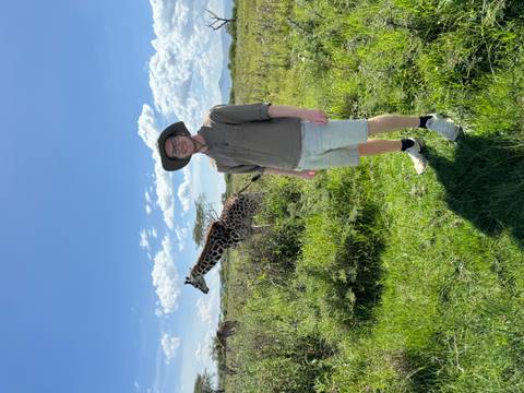       Traveler stands on grassy plain while a giraffe grazes nearby under a clear sky.
  