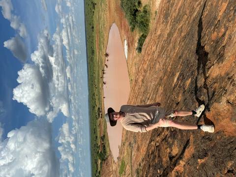       Traveler stands atop a rocky ridge overlooking a clay-colored watering hole where buffalo cool off under dramatic clouds.
  