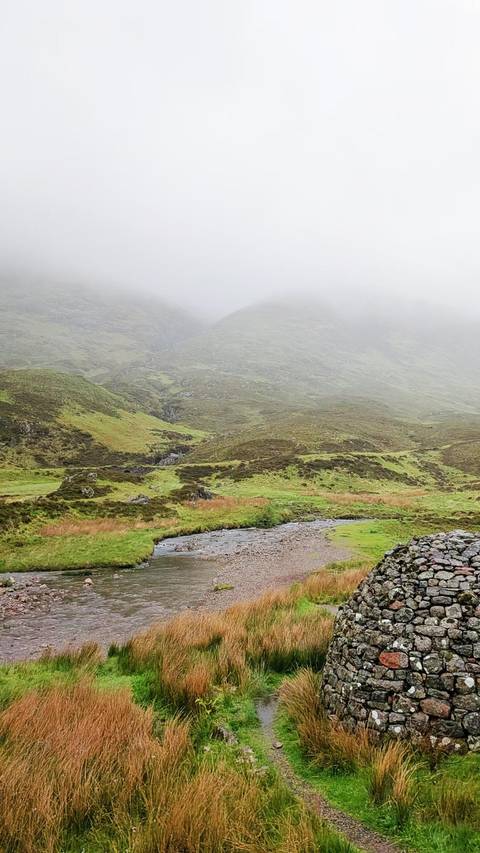       Misty green valley with a stream winding through grassy hills under low clouds.
  