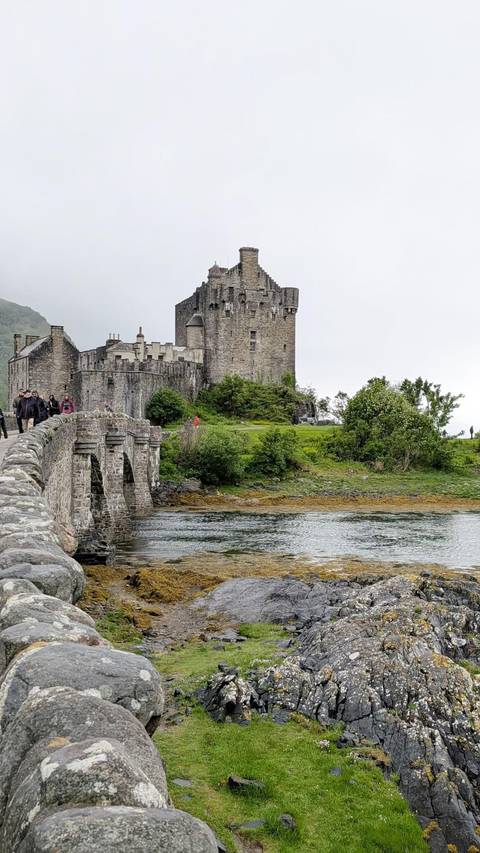       Stone bridge leading to a historic castle on a loch shore with visitors walking across.
  