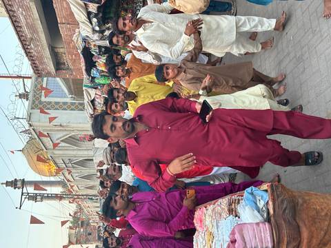       Crowded marketplace street with a man in a red kurta striding toward the camera surrounded by shoppers and colorful stalls.
  