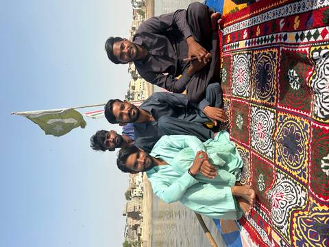       Four young men sitting cross-legged on a patterned carpet atop a boat on a river, a Pakistani flag fluttering behind them.
  