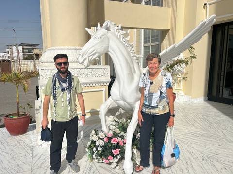       Two travelers posing beside a life-size white horse statue outside a marble-floored building entrance.
  