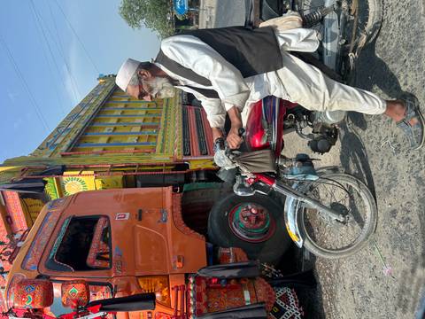       Elderly man with white beard riding a red motorbike beside colorfully painted Pakistani freight trucks on a sunny road.
  