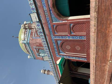       Brick and tile mausoleum with green dome and patterned facade under a bright blue sky with a few birds flying.
  