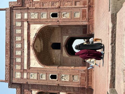       Women in traditional dress and a child walk through a grand red sandstone arched gateway richly decorated with geometric carvings.
  