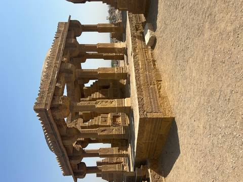       Intricately carved golden sandstone tomb pavilion standing under a clear blue sky in a desert landscape.
  