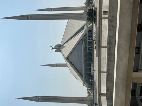       Iconic white Faisal Mosque with angular roof and twin minarets viewed across an open plaza on a hazy day.
  