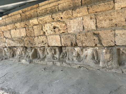       Eroded stone wall base featuring ancient carved reliefs of seated figures inside an archaeological site.
  