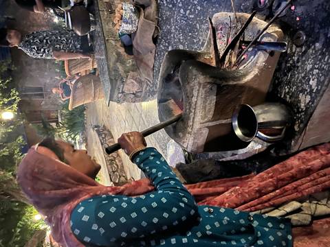       Local woman in traditional dress grinding spices with large pestle beside outdoor hearth at dusk.
  