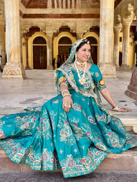       Woman dressed in ornate turquoise sari poses gracefully inside palace courtyard.
  
