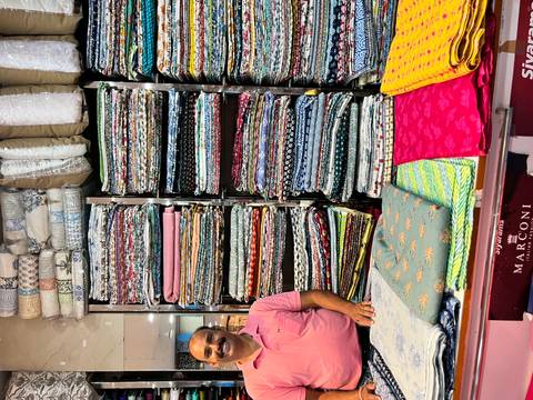       Colorful stacks of fabric bolts line walls of textile shop with smiling shopkeeper.
  