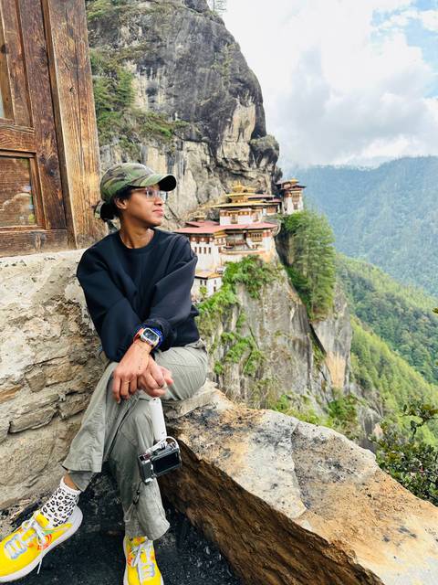       Woman seated on a rocky ledge gazes at Bhutan's cliff-hugging Tiger's Nest Monastery surrounded by forested mountains.
  