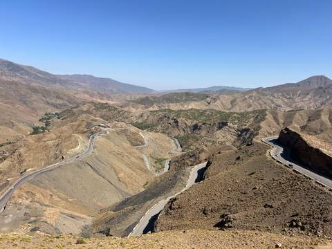       Sweeping aerial view of winding asphalt road snaking through barren brown mountains under a clear blue sky.
  