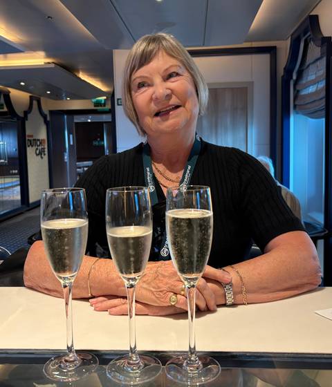       Smiling woman seated at a table with three flutes of sparkling wine on a cruise ship lounge.
  