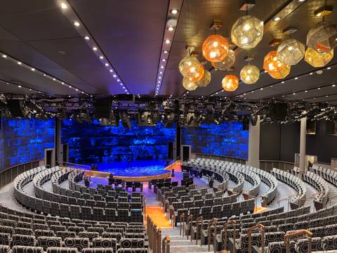       Modern circular theatre auditorium on a cruise ship with blue illuminated stage and decorative ceiling lights.
  
