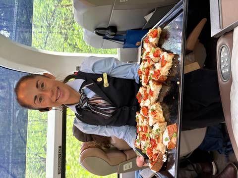       Smiling attendant on panoramic train carriage presenting a tray of gourmet bruschetta snacks.
  
