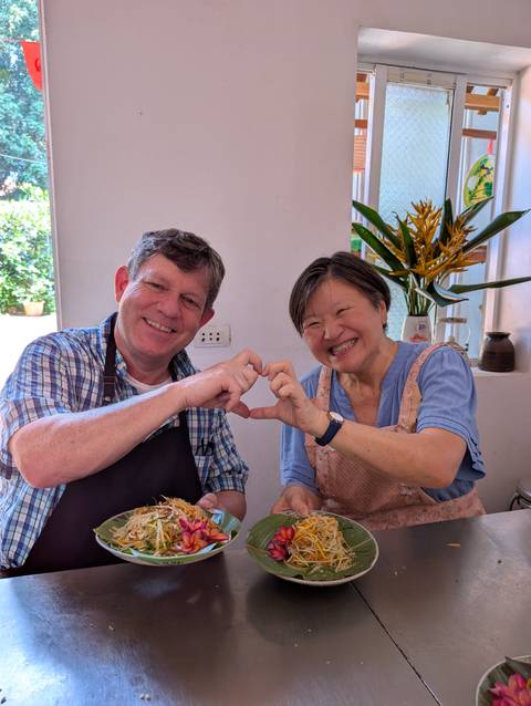       A couple seated at a table form a heart shape with their hands and laugh together.
  