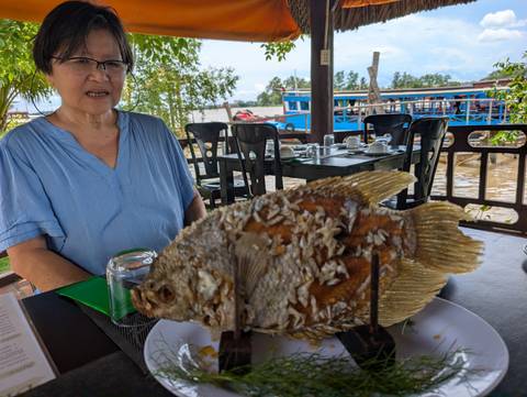       A whole fried fish served upright on a platter at a riverside restaurant with a diner looking on.
  