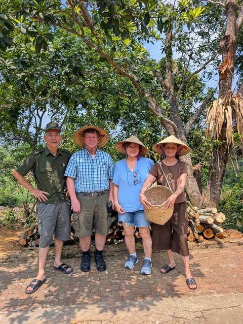       Four people wearing sun hats pose with a forest ranger among trees and stacked firewood.
  
