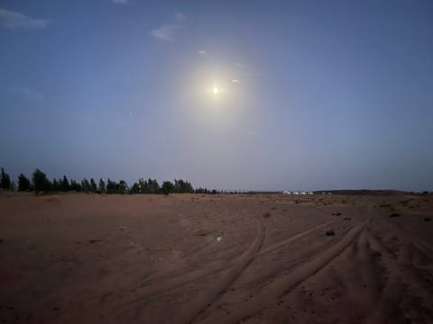       Moonlit Sahara desert landscape with faint camp tents visible on the horizon
  