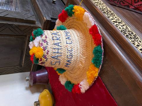       Hand-woven straw hat decorated with multicolored pompoms labeled 'Sahara Magic Camp'
  